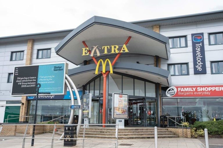 A McDonald's restaurant located in a multi-business building within Motorway Service Areas, with an adjacent Travelodge hotel and family shopper store.