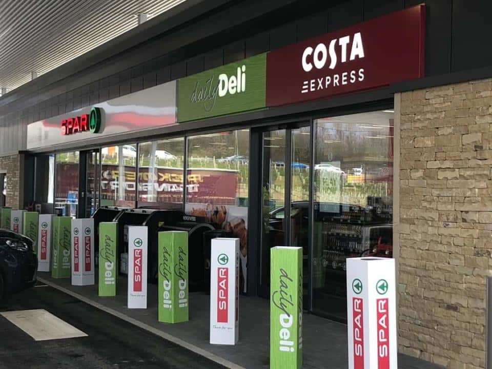 A motorway petrol station entrance with spar and costa express branding, flanked by promotional signs.