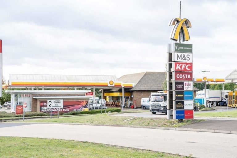 A Motorway Service Area with multiple franchises including a gas station, KFC, M&S, and Costa Coffee, along with signage displaying brand logos and fuel prices.