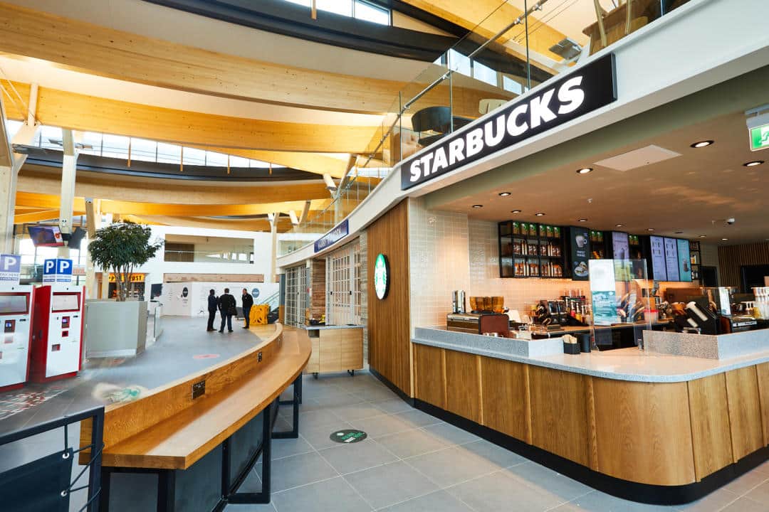 Modern Starbucks coffee shop located in a spacious interior with natural light and wood accents, offering food and drink options near the motorway.