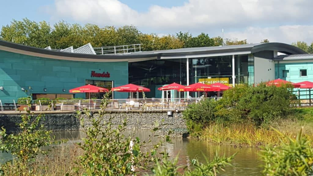 A lakeside restaurant with outdoor seating and red umbrellas displaying the nando's logo on a sunny day.