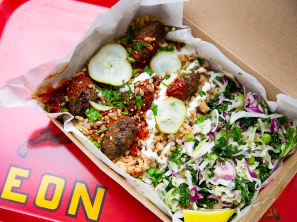 A takeaway box filled with rice, meatballs, fresh herbs, slaw, and sliced cucumbers, served on a red surface.