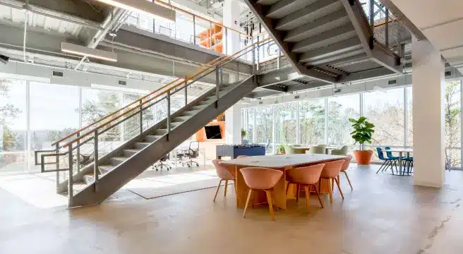 Modern office with open layout featuring a steel staircase, large windows, concrete floor, and stylish furniture including peach chairs and a wooden table; located near a motorway service area for extra convenience.