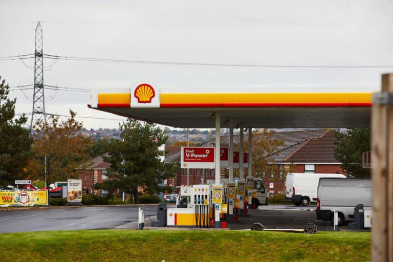 A shell gas station with a bright yellow and red canopy, fuel pumps, and a truck parked nearby, set against a motorway service area backdrop.