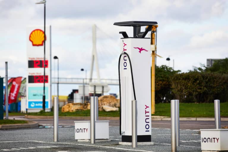 An ionity electric vehicle charging station with multiple chargers, located near a motorway service area with a shell gas station and price sign in the background.