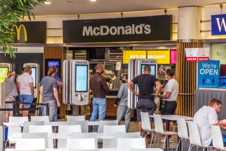 Customers ordering food at self-service kiosks in a busy McDonald's restaurant located in a motorway service area, with seating area in the foreground.
