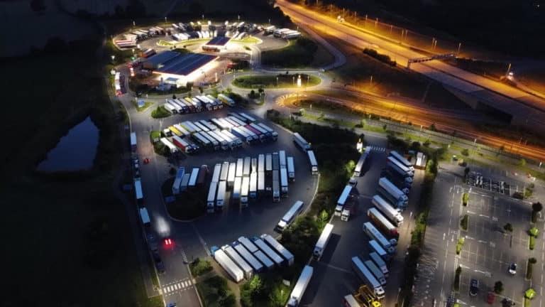 Aerial night view of a motorway service area with rows of trucks parked adjacent to a brightly lit extra services station and roundabout.