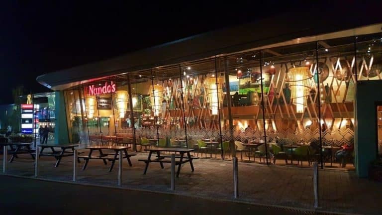 Exterior view of a Nando's restaurant at a motorway service area at night, illuminated and visible through large glass windows with decorative patterns.