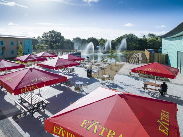 Outdoor café at a motorway service area with red umbrellas, tables, and people sitting near a pond with fountains on a sunny day.