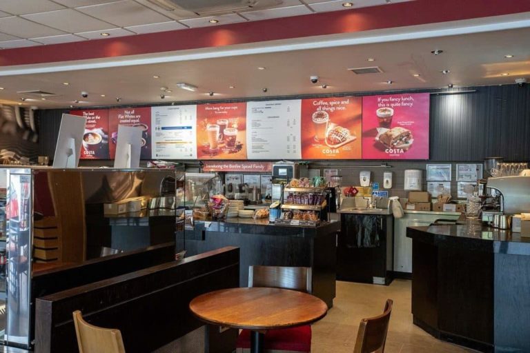 Interior of a Costa Coffee shop at a motorway service area, showcasing the counter, menu boards, and seating area.