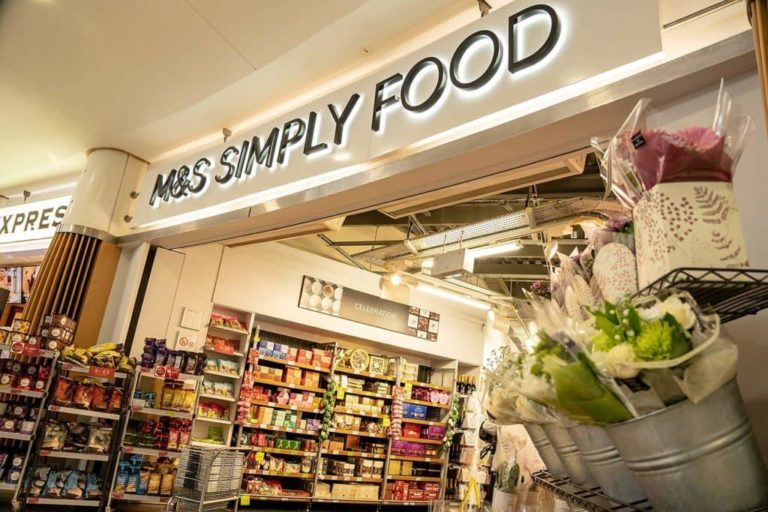 Neon-lit sign "m&s simply food" above a grocery store entrance at a motorway service area, featuring a flower display and shelves stocked with assorted products.