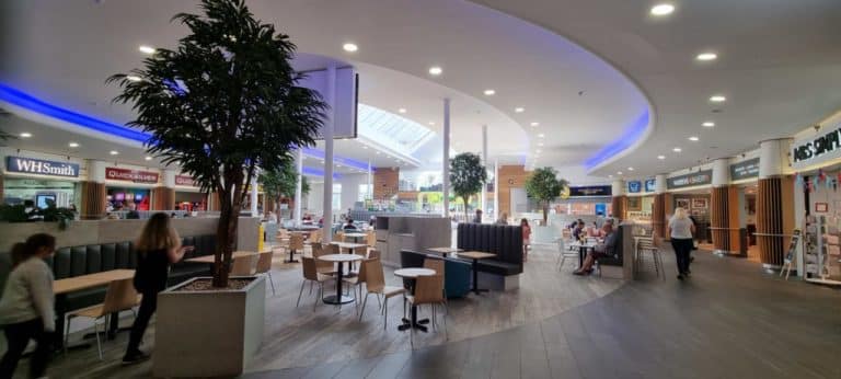 Interior of a modern motorway service area food court with patrons at tables, shops in the background, and a palm tree.