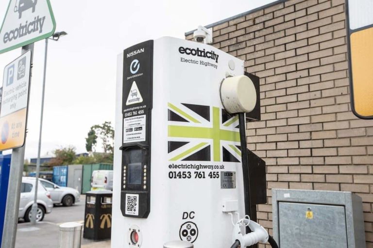 Electric car charging station by Ecotricity with various adapter options, displayed outside at a motorway service area against a building.