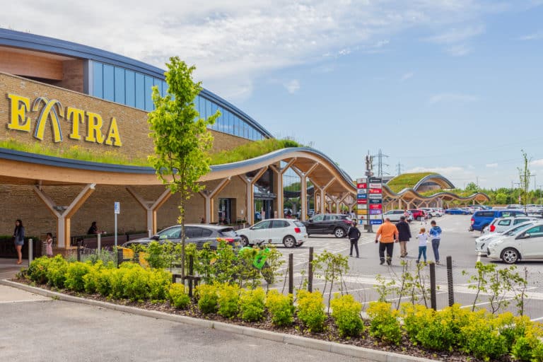 Exterior view of a modern extra supermarket with arched wooden canopy, featuring parked cars and shoppers on a sunny day.