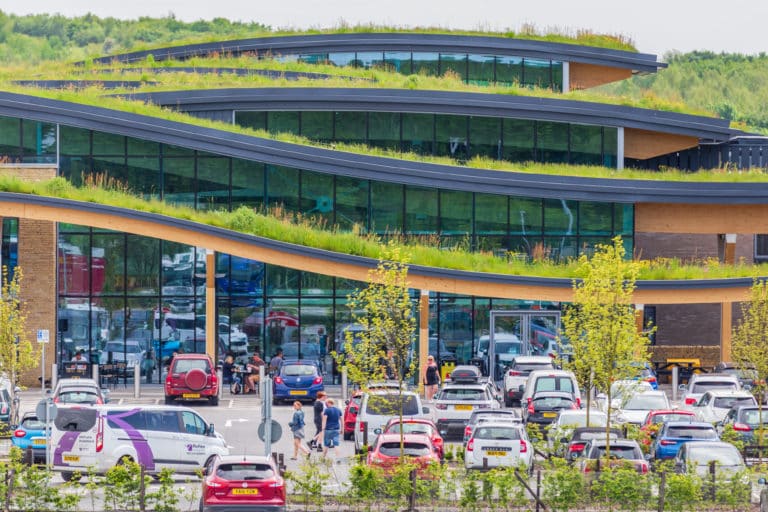 Modern building with curved, layered architecture and green roof, surrounded by busy parking lot with cars and pedestrians.