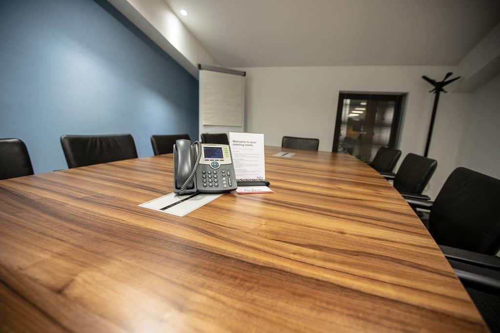 Modern conference room with a large wooden table featuring a telephone and papers, black chairs, and a blue wall.