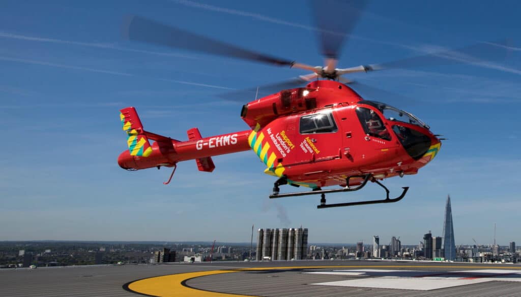 Red emergency helicopter in flight over a city, with 'London's Air Ambulance' markings visible on its side and a tall skyline below.